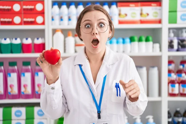 Young Caucasian Woman Working Pharmacy Drugstore Holding Red Heart Scared — Zdjęcie stockowe