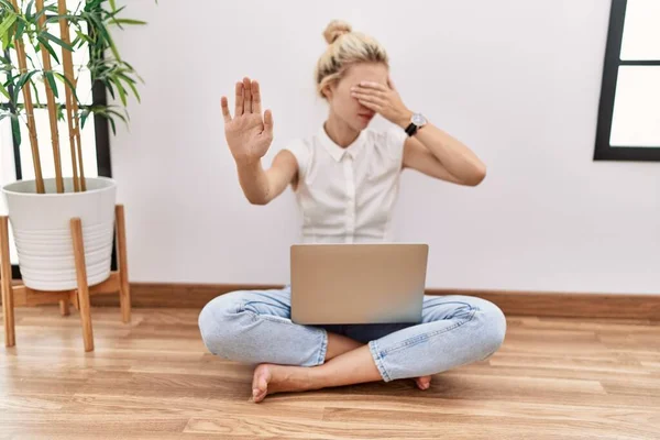 Young blonde woman using computer laptop sitting on the floor at the living room covering eyes with hands and doing stop gesture with sad and fear expression. embarrassed and negative concept.