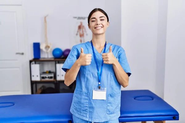 理学療法士を身に着けている若いヒスパニック系の女性は 手で積極的なジェスチャーを行う診療所の成功サインに立って均一性を主張します 親指アップ笑顔と幸せ 陽気な表情と勝者のジェスチャー — ストック写真