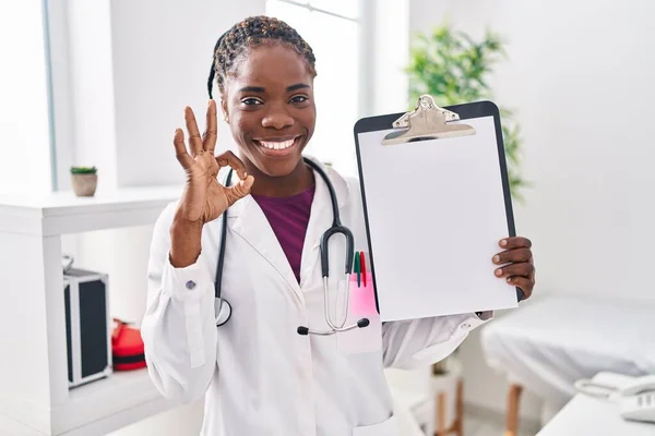 Beautiful black doctor woman holding clipboard doing ok sign with fingers, smiling friendly gesturing excellent symbol