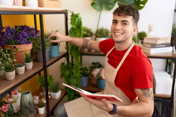Young hispanic man florist reading book holding plant of shelving at flower shop