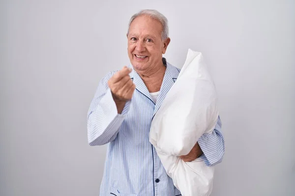 Senior man with grey hair wearing pijama hugging pillow doing money gesture with hands, asking for salary payment, millionaire business