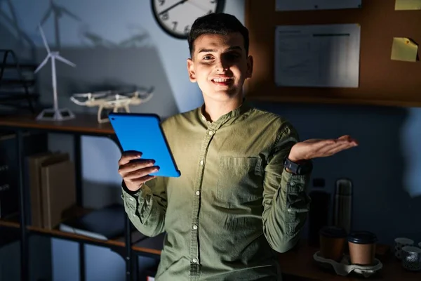 Non binary person using touchpad device at night smiling cheerful presenting and pointing with palm of hand looking at the camera.