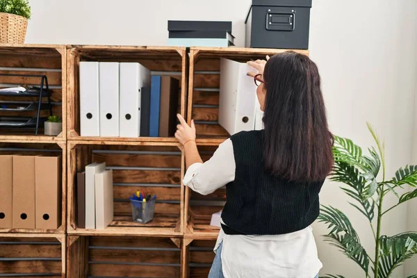 Young beautiful hispanic woman business worker holding documents of shelving at office