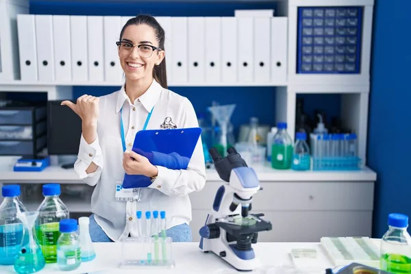 Young brunette woman working at scientist laboratory pointing to the back behind with hand and thumbs up, smiling confident
