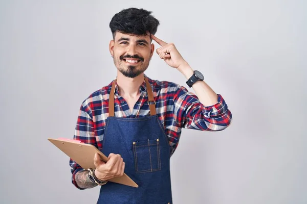 Young hispanic man with beard wearing waiter apron holding clipboard smiling pointing to head with one finger, great idea or thought, good memory