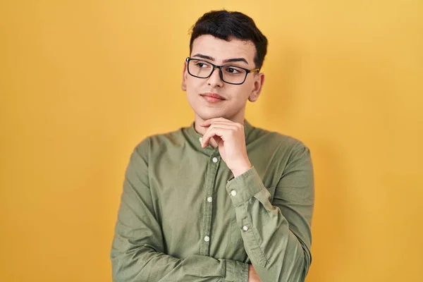Non binary person standing over yellow background with hand on chin thinking about question, pensive expression. smiling with thoughtful face. doubt concept.