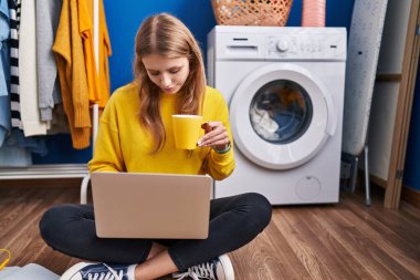 Young blonde woman using laptop drinking coffee waiting for washing machine at laundry room