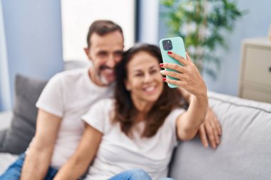 Orta yaşlı erkek ve kadın çift evdeki kanepede oturan akıllı telefonun yanında selfie çekiyorlar.