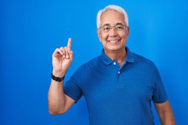 Middle age man with grey hair standing over blue background showing and pointing up with finger number one while smiling confident and happy. 
