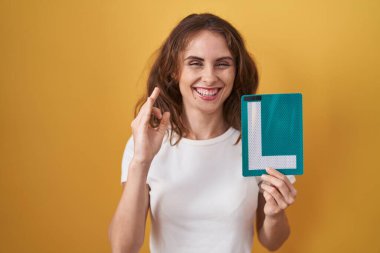 Beautiful brunette woman holding l sign for new driver doing ok sign with fingers, smiling friendly gesturing excellent symbol 