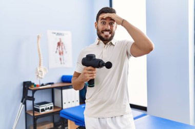 Handsome hispanic man holding therapy massage gun at physiotherapy center stressed and frustrated with hand on head, surprised and angry face 