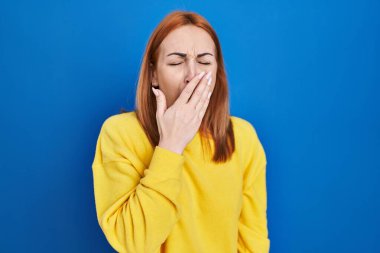 Young woman standing over blue background bored yawning tired covering mouth with hand. restless and sleepiness. 