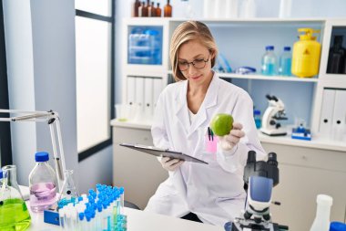 Young blonde woman scientist holding apple reading report at laboratory
