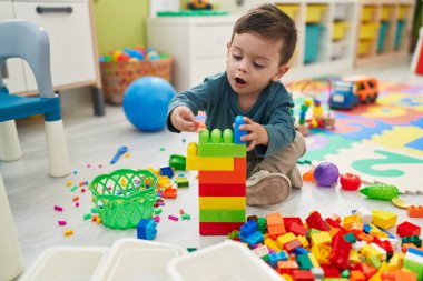 Adorable hispanic boy playing with construction blocks sitting on floor at kindergarten