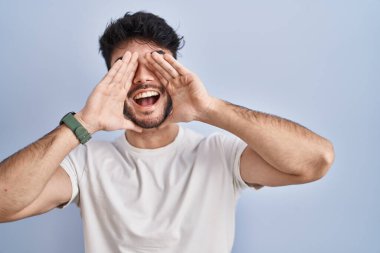 Hispanic man with beard standing over white background smiling cheerful playing peek a boo with hands showing face. surprised and exited 