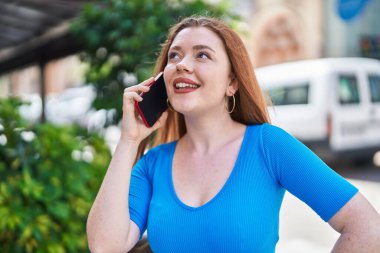 Genç kızıl saçlı, gülümseyen, kendine güvenen, caddedeki akıllı telefondan konuşan kadın.