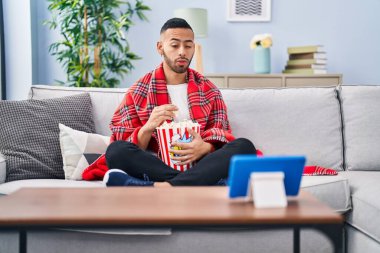 Young hispanic man eating popcorn watching movie on tablet device making fish face with mouth and squinting eyes, crazy and comical. 