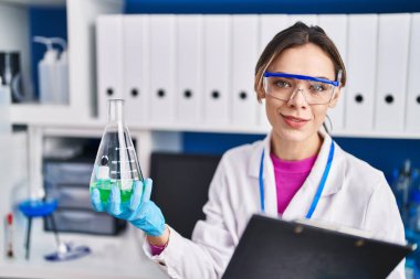Young beautiful hispanic woman scientist reading report holding test tube at laboratory