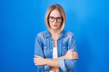 Mavi arka planda duran beyaz bir kadın şüpheci ve gergin, yüzünde çapraz kollarla onaylamayan bir ifade. negatif kişi. 