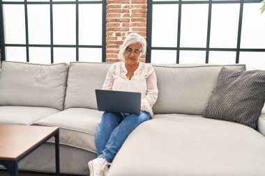 Orta yaşlı bir kadın evdeki koltukta dizüstü bilgisayar kullanıyor.