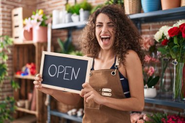 Hispanic woman with curly hair working at florist holding open sign smiling and laughing hard out loud because funny crazy joke. 