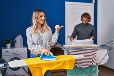 Couple ironing clothes at laundry room pointing thumb up to the side smiling happy with open mouth 