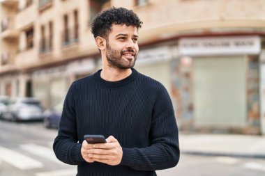 Genç Arap adam sokakta akıllı telefon kullanarak kendinden emin gülümsüyor.