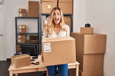 Young blonde woman working at small business ecommerce holding big box celebrating crazy and amazed for success with open eyes screaming excited. 