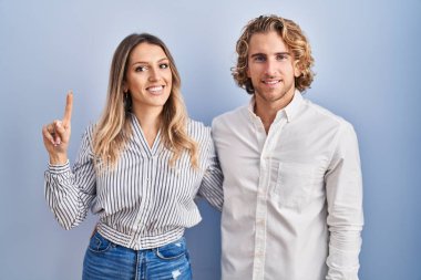 Young couple standing over blue background showing and pointing up with finger number one while smiling confident and happy. 