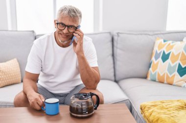 Middle age grey-haired man drinking coffee and talking on the smartphone at home