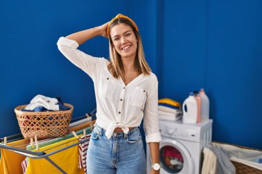 Çamaşır odasındaki genç sarışın kadın gülümsüyor. Kendine güvenen, el hareketleriyle saçlara dokunan, çekici ve modaya uygun poz veren. 