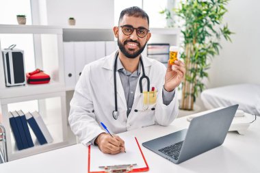 Young hispanic man doctor holding pills bottle writing on document at clinic