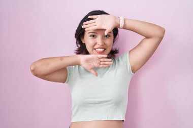 Hispanic young woman standing over pink background smiling cheerful playing peek a boo with hands showing face. surprised and exited 