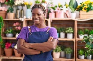 Afro-Amerikan kadın çiçekçi gülümsüyor. Çiçekçiye kollarını kavuşturmuş duruyor.