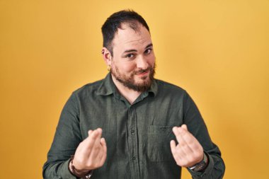 Plus size hispanic man with beard standing over yellow background doing money gesture with hands, asking for salary payment, millionaire business 