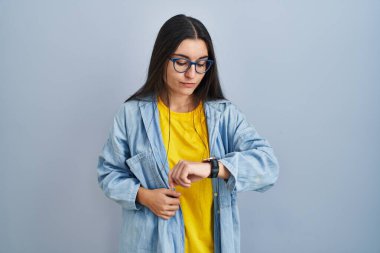 Mavi arka planda duran genç İspanyol kadın saati kontrol ediyor, rahat ve kendinden emin. 