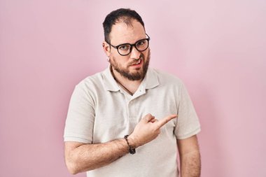 Plus size hispanic man with beard standing over pink background pointing aside worried and nervous with forefinger, concerned and surprised expression 
