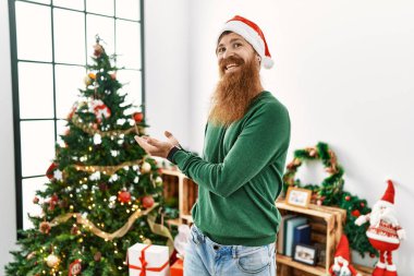Redhead man with long beard wearing christmas hat by christmas tree pointing aside with hands open palms showing copy space, presenting advertisement smiling excited happy 