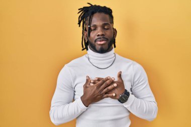 Sarı arka planda balıkçı yaka kazak giyen Afrikalı bir adam elleri göğsünde, gözleri kapalı ve yüzünde minnet dolu bir jest ile gülümsüyor. sağlık kavramı. 