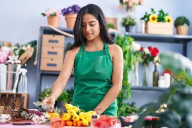 Young hispanic woman florist make bouquet of flowers at florist shop