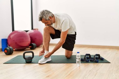Middle age grey-haired man tying shoe at sport center