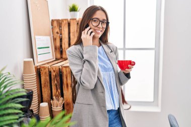Young beautiful hispanic woman business worker talking on smartphone drinking coffee at office
