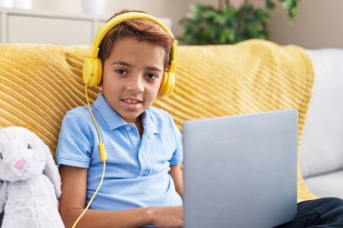 Adorable hispanic boy using laptop and headphones sitting on sofa at home