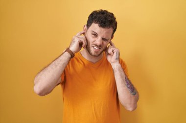 Sarı arka planda dikilen İspanyol genç adam yüksek sesli müziğin gürültüsü için parmaklarıyla kulaklarını kapatıyor. sağır kavramı. 