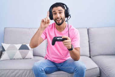 Hispanic young man playing video game holding controller sitting on the sofa pointing finger up with successful idea. exited and happy. number one. 