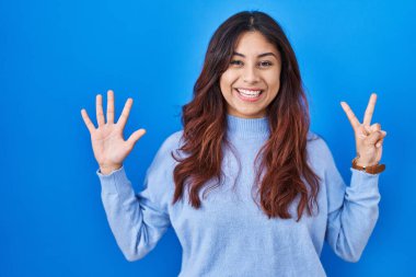 Hispanic young woman standing over blue background showing and pointing up with fingers number seven while smiling confident and happy. 