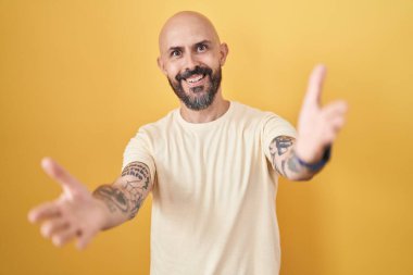 Hispanic man with tattoos standing over yellow background looking at the camera smiling with open arms for hug. cheerful expression embracing happiness. 