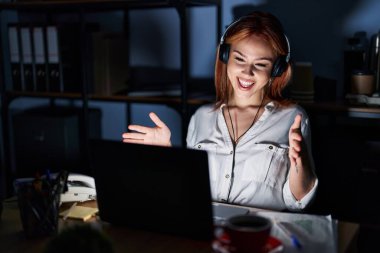 Young caucasian woman working at the office at night looking at the camera smiling with open arms for hug. cheerful expression embracing happiness. 