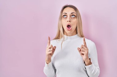 Young blonde woman wearing white sweater over pink background amazed and surprised looking up and pointing with fingers and raised arms. 